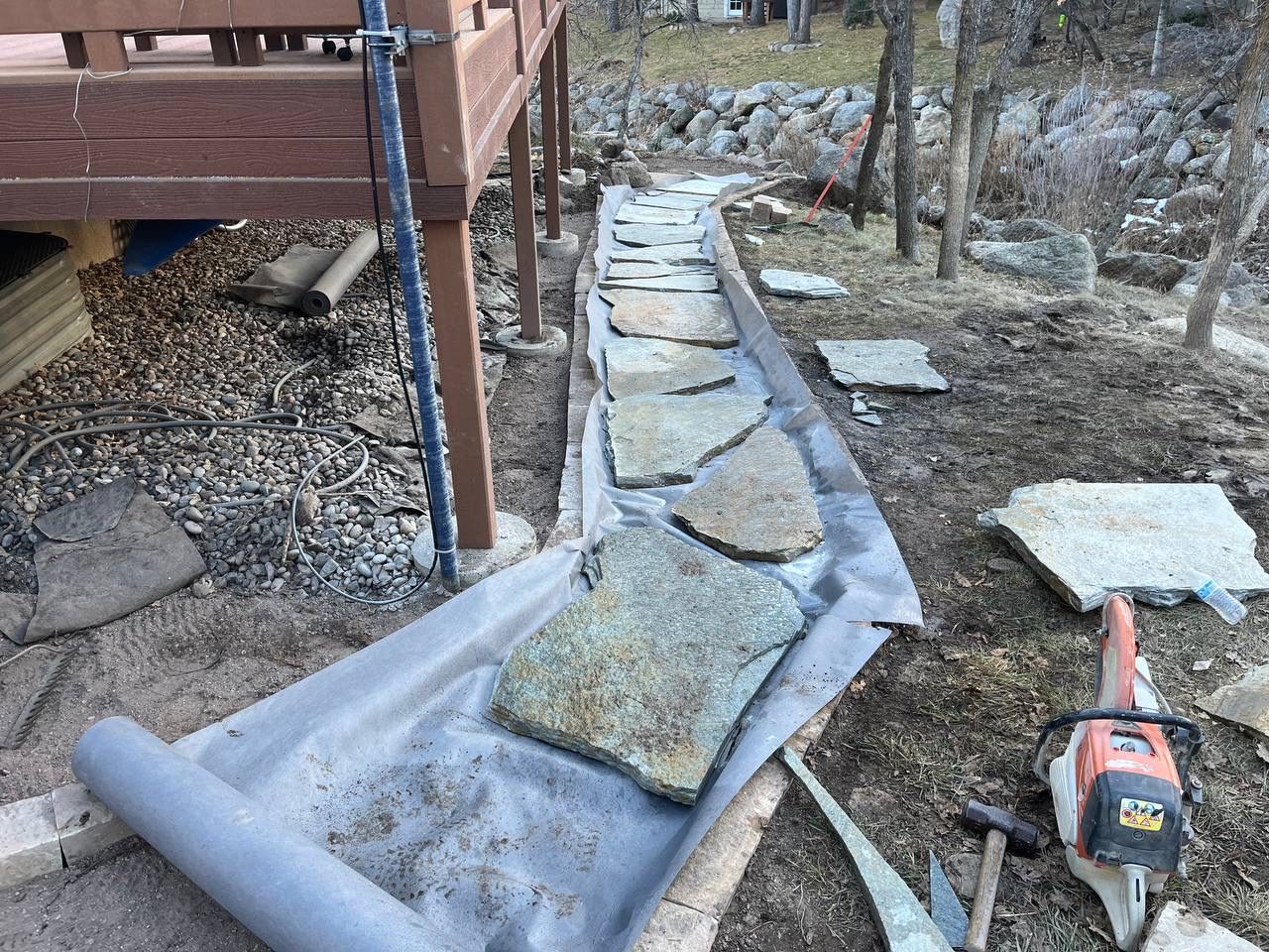 Stone walkway under construction beside a deck, with gravel, tarps, and tools in a yard area