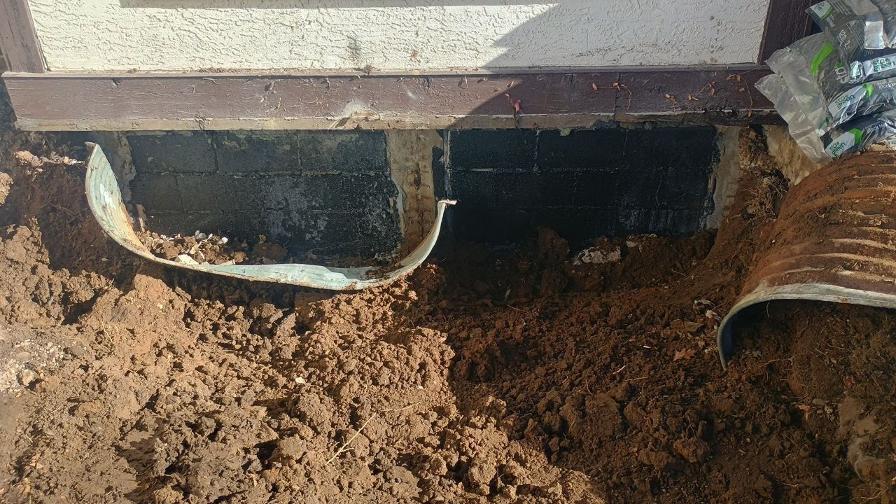 Two white and rusted pipes protrude from a wall into muddy soil