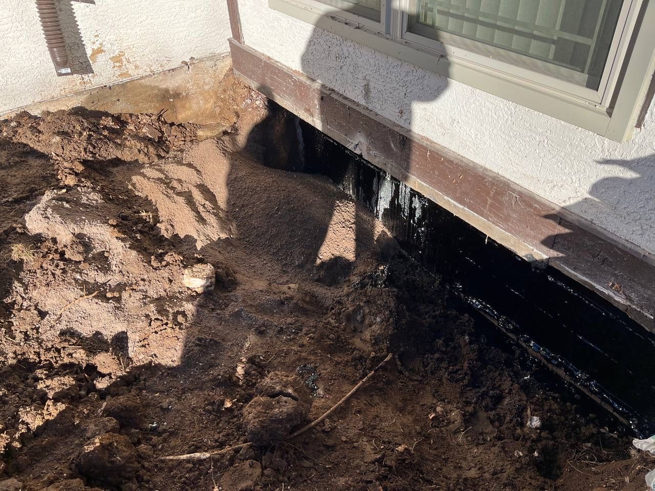 Eroded soil beside a house foundation with exposed black waterproofing under a window