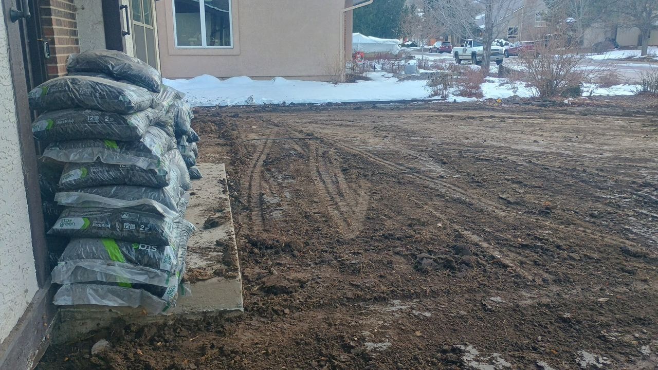 Stacked bags of soil beside a muddy yard near a building with patches of snow.
