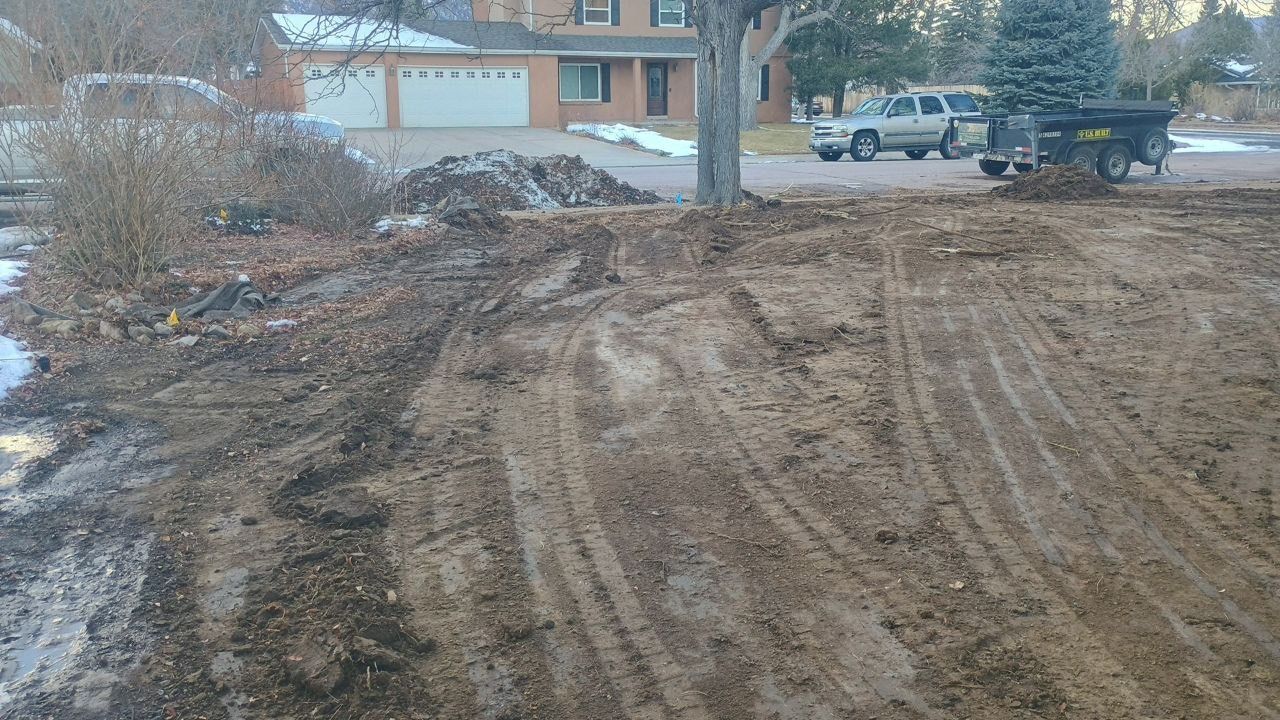 Muddy roadside area with tire tracks, snow patches, and parked vehicles near houses