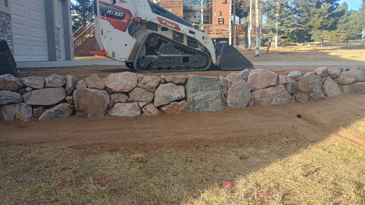 Skid steer loader perched on a stone retaining wall beside a house and lawn under construction.