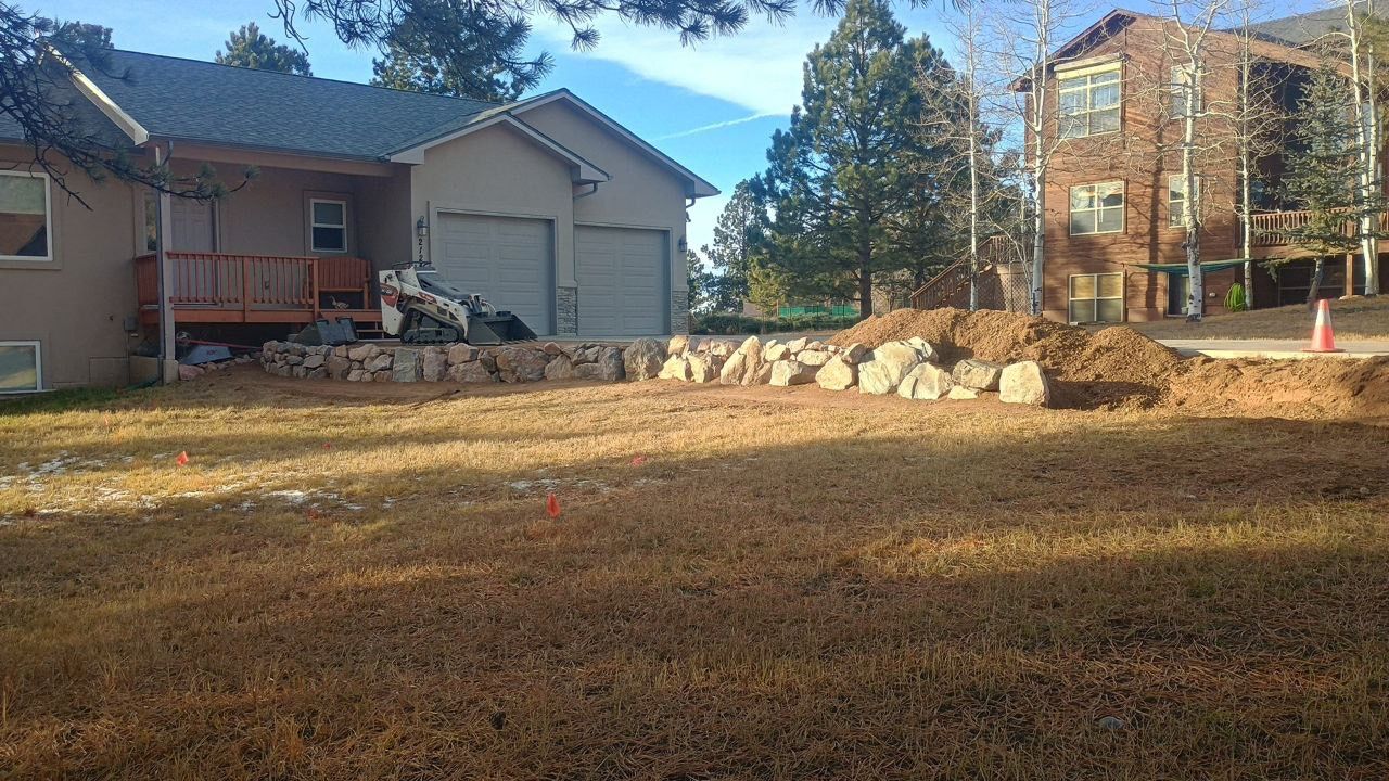 Residential yard with brown grass, a woodpile, and houses under a blue sky
