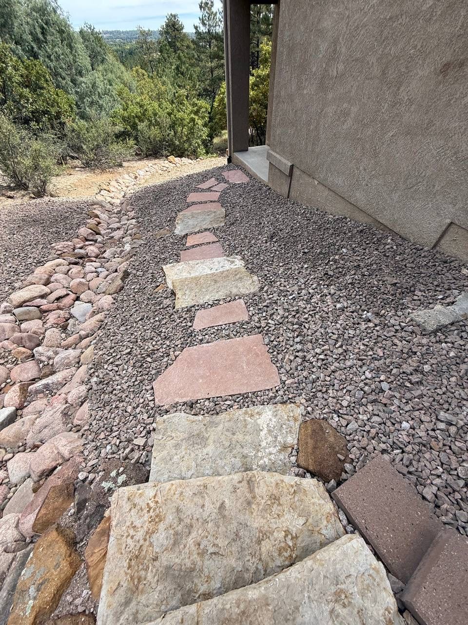 Stone garden path beside a stucco wall, bordered by gravel and pink pavers leading toward trees