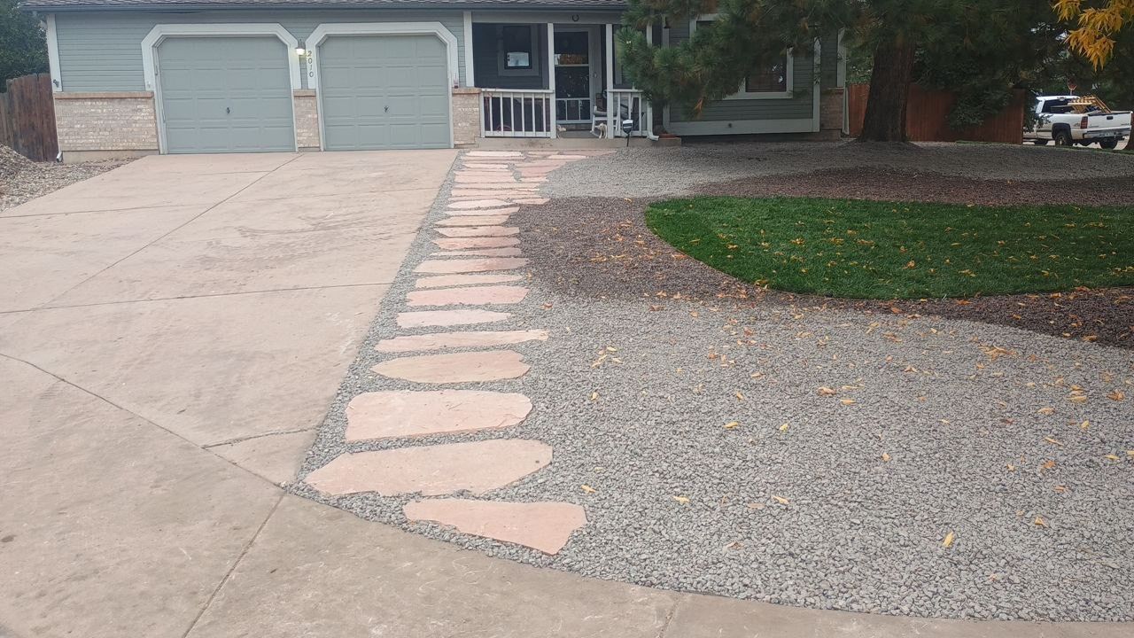 Front yard with a curved stone walkway, gravel driveway, two-car garage, and trimmed lawn.