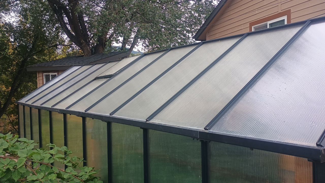 Long greenhouse with a slanted translucent roof beside a brick house and garden greenery