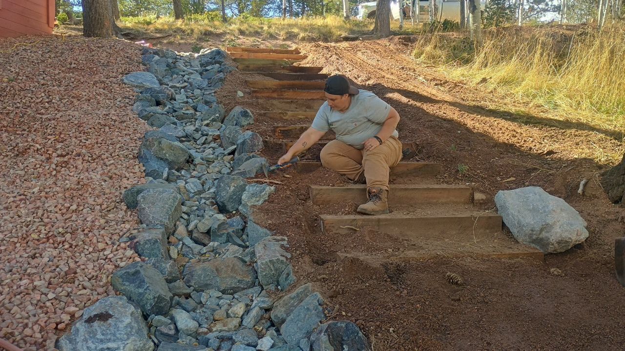 Person crouching beside a rocky drainage channel on a dirt hillside, placing stones in a landscaped area