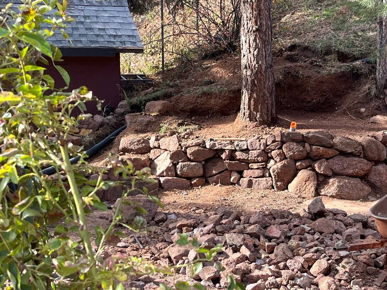 Stone retaining wall and red shed in a wooded yard with a large tree and rocky ground