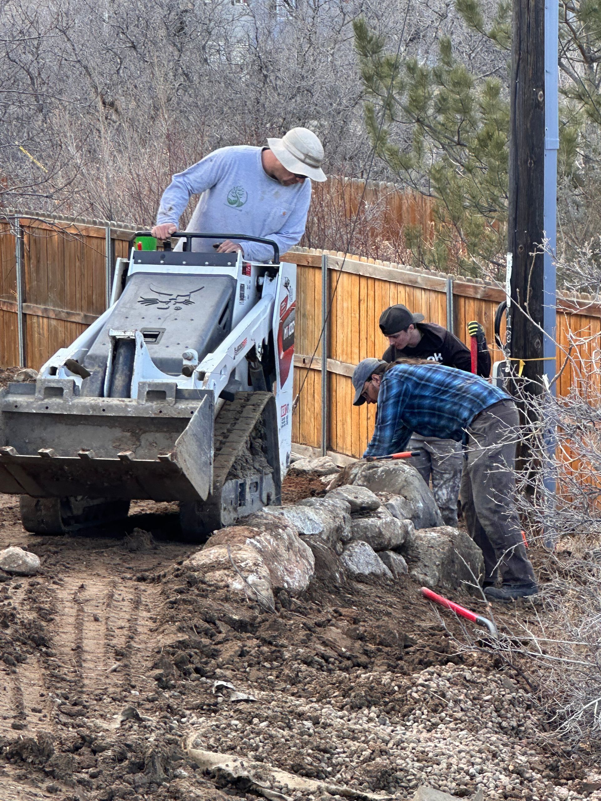 Workers guide a skid steer over rocks along a muddy slope near a fence.