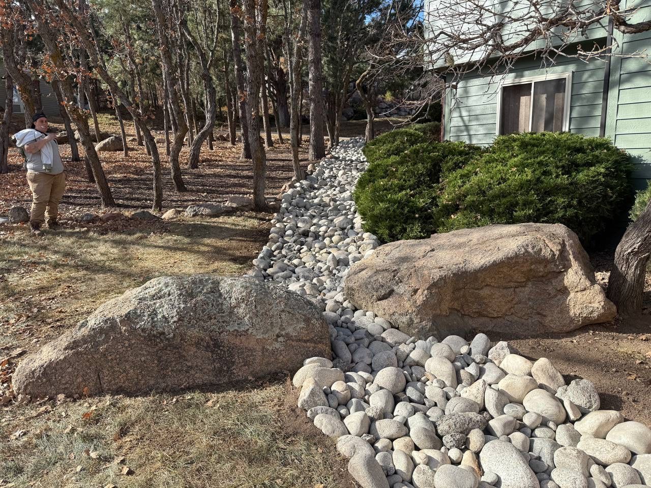 Landscaped yard with large rocks, white gravel, shrubs, and a small green building in the background