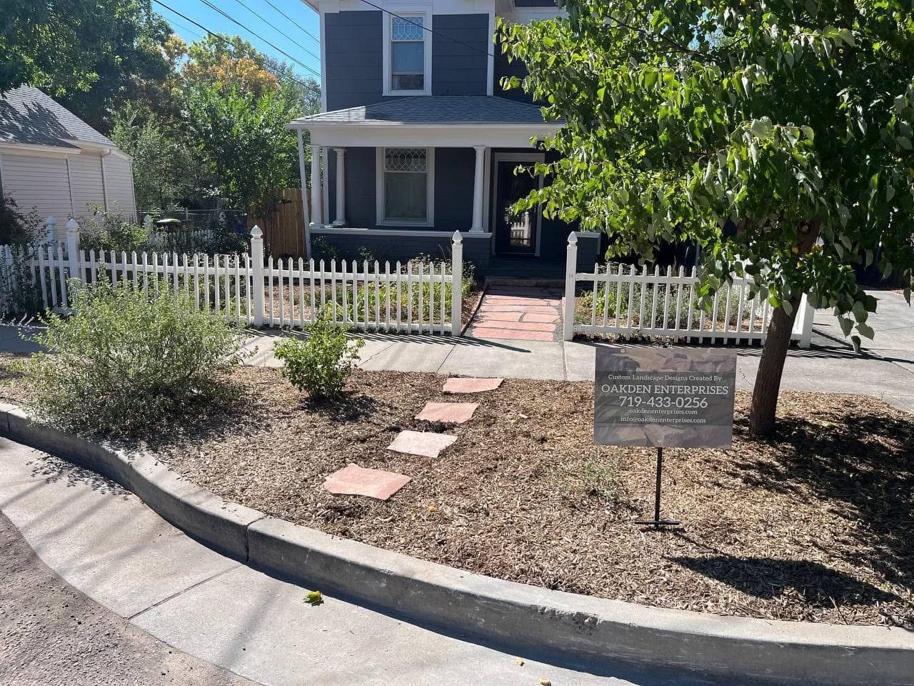 Two-story house with white picket fence, front walkway, and yard sign beside a tree and shrubs.