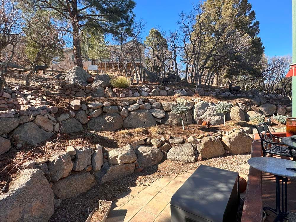 Stone retaining walls and patio overlook a hillside with trees and distant buildings on a sunny day.