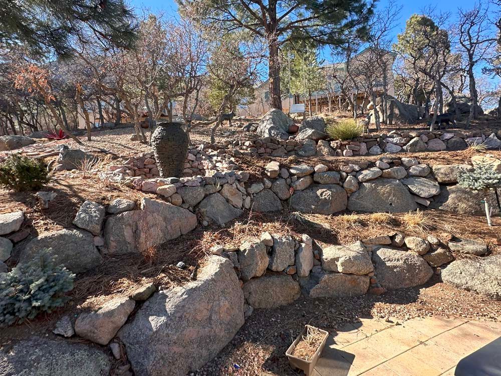 Rocky hillside with stone retaining walls, scattered plants, and a vase, under a blue sky.