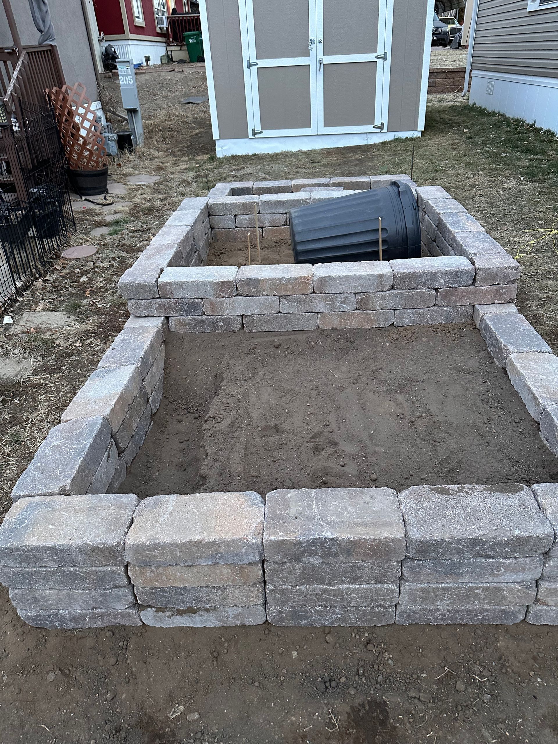Raised rectangular stone fire pit in a backyard patio area, with a gravel-filled center and a white shed behind.