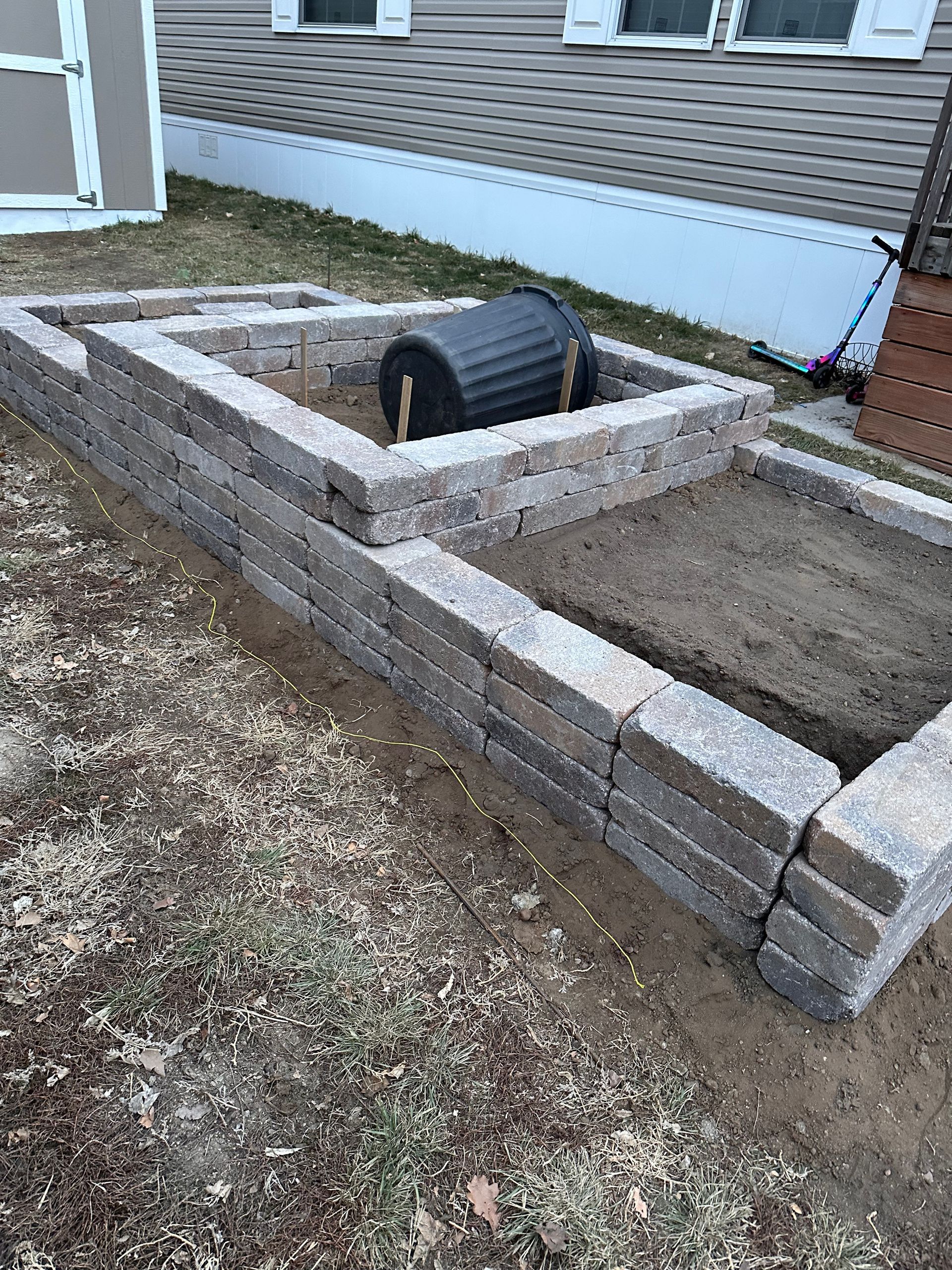 Raised brick garden bed with a black pipe or wheel in the center beside a house