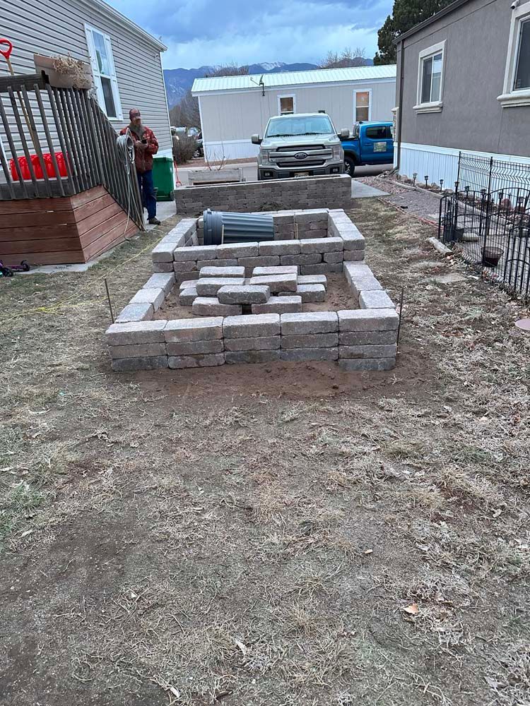 Raised garden beds in a yard, with a person standing nearby, surrounded by brown dirt and homes in the background.