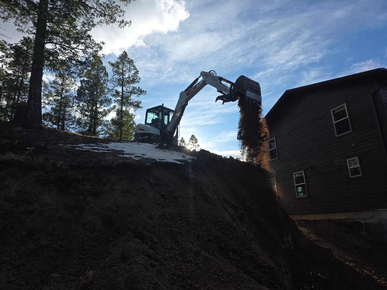 Excavator dumping soil onto a large pile beside a building at sunset