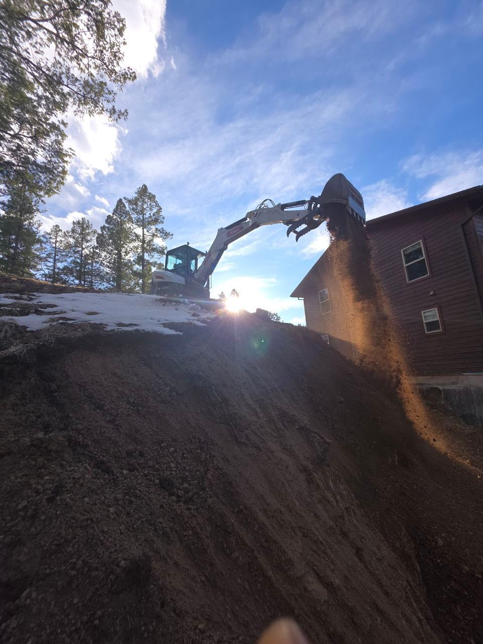 Excavator dumping dirt beside a brick building on a snowy slope at sunset