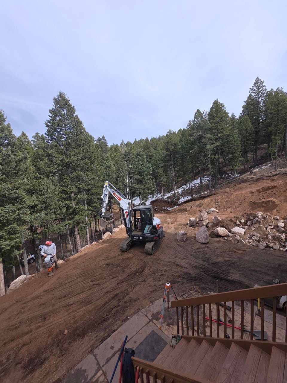 Backhoe excavator working on a dirt slope in a wooded area, with a person nearby and a deck in foreground