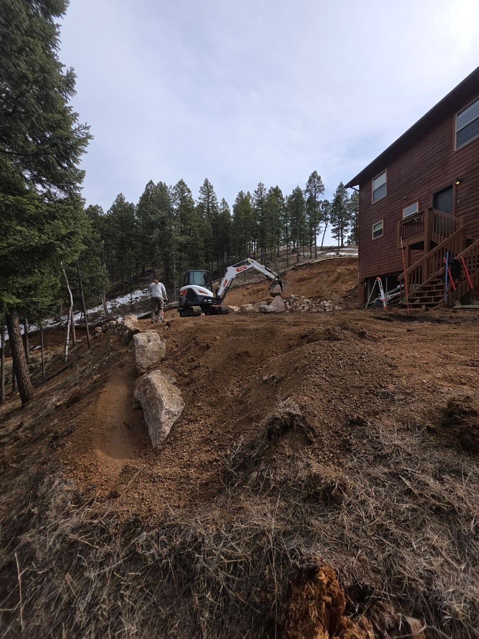 Landslide debris beside a brick building and wooded hillside, with a muddy slope and downed trees.
