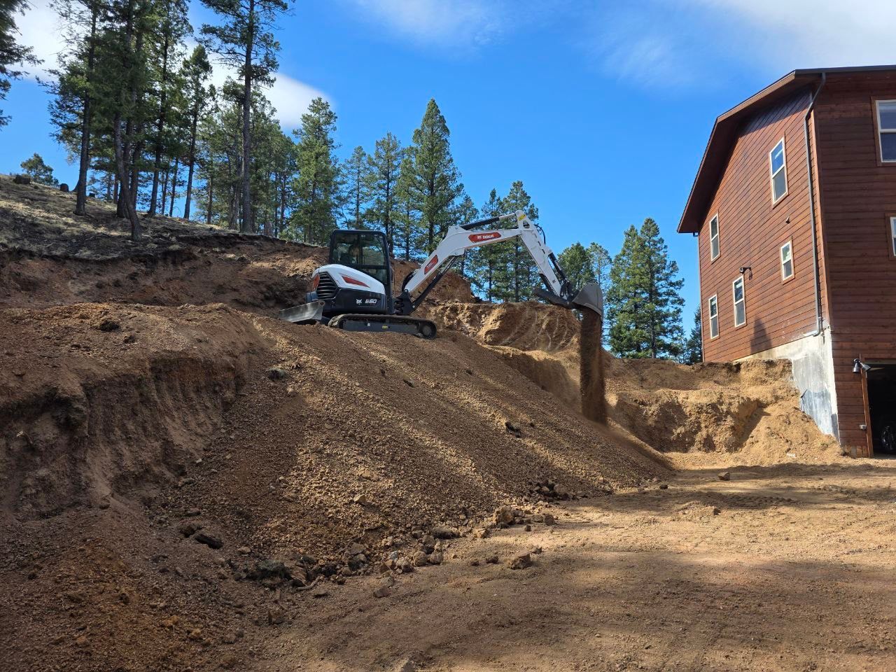 Excavator grading a dirt hillside beside a red wooden building in a wooded area