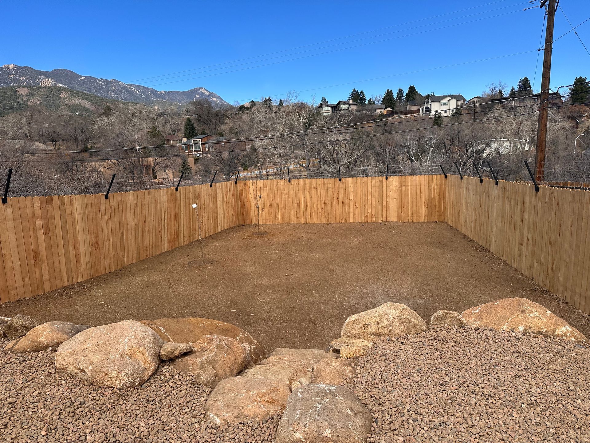 Wooden-fenced dirt yard with large rocks, backed by hills and a clear blue sky