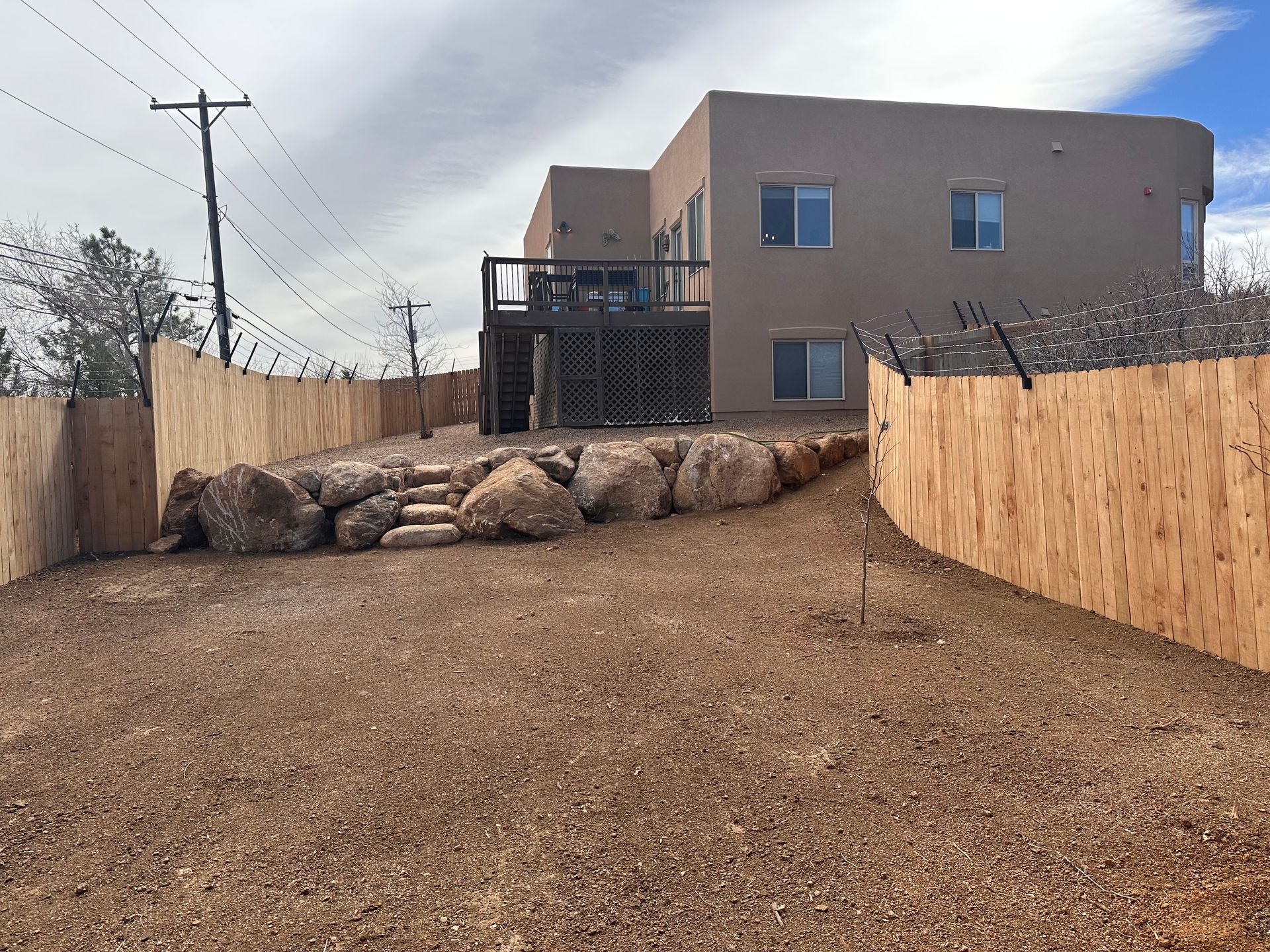 Backyard with gravel path, tan privacy walls, stacked rocks, and a two-story beige house under cloudy sky