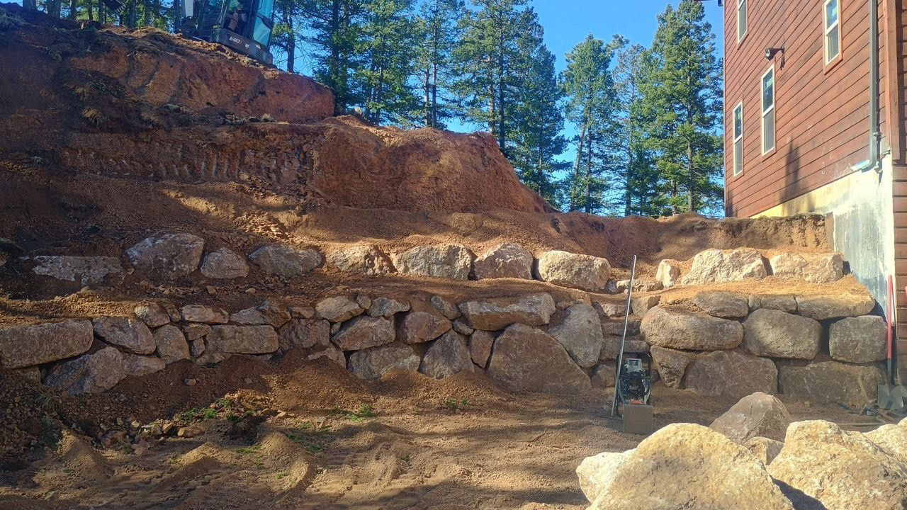 Rock retaining wall on a dirt slope beside a brick building, with pine trees and blue sky above.