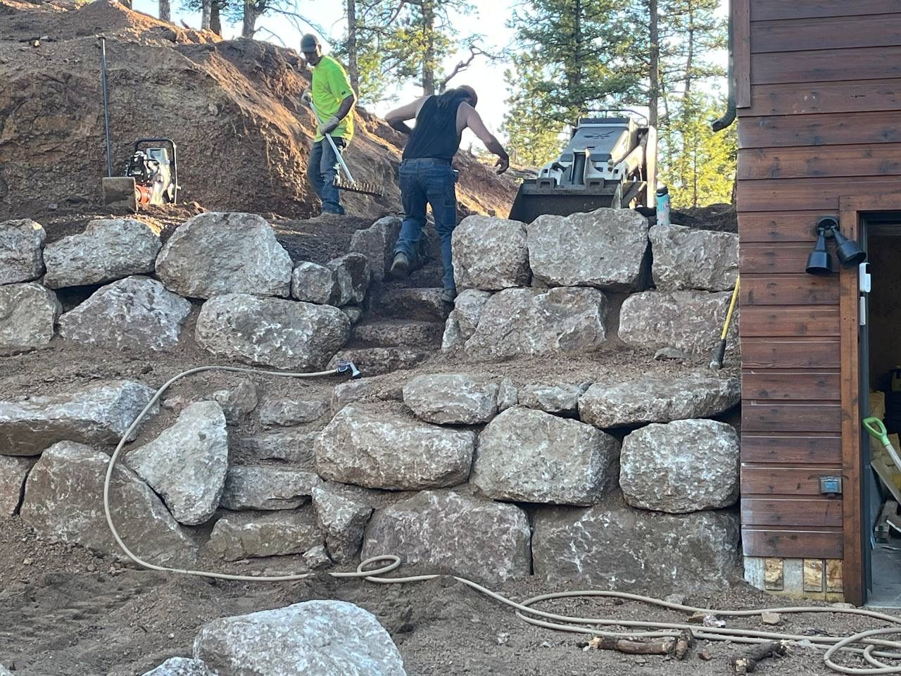 Two workers stacking large stones to build a retaining wall beside a wooden house in a wooded yard