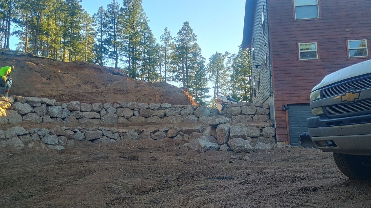 Rock retaining wall beside a red building, with trees on a hillside and a truck at right.