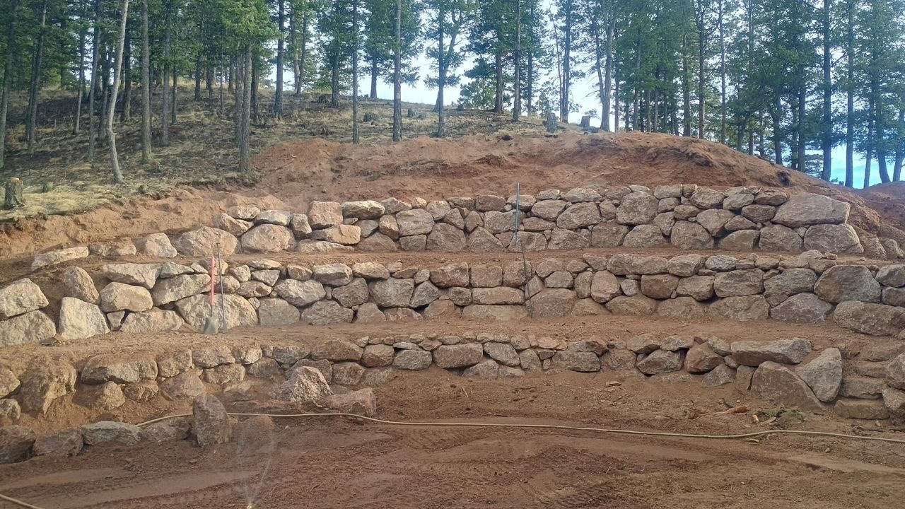 Terraced stone retaining wall on a wooded hillside with exposed earth above and dirt below