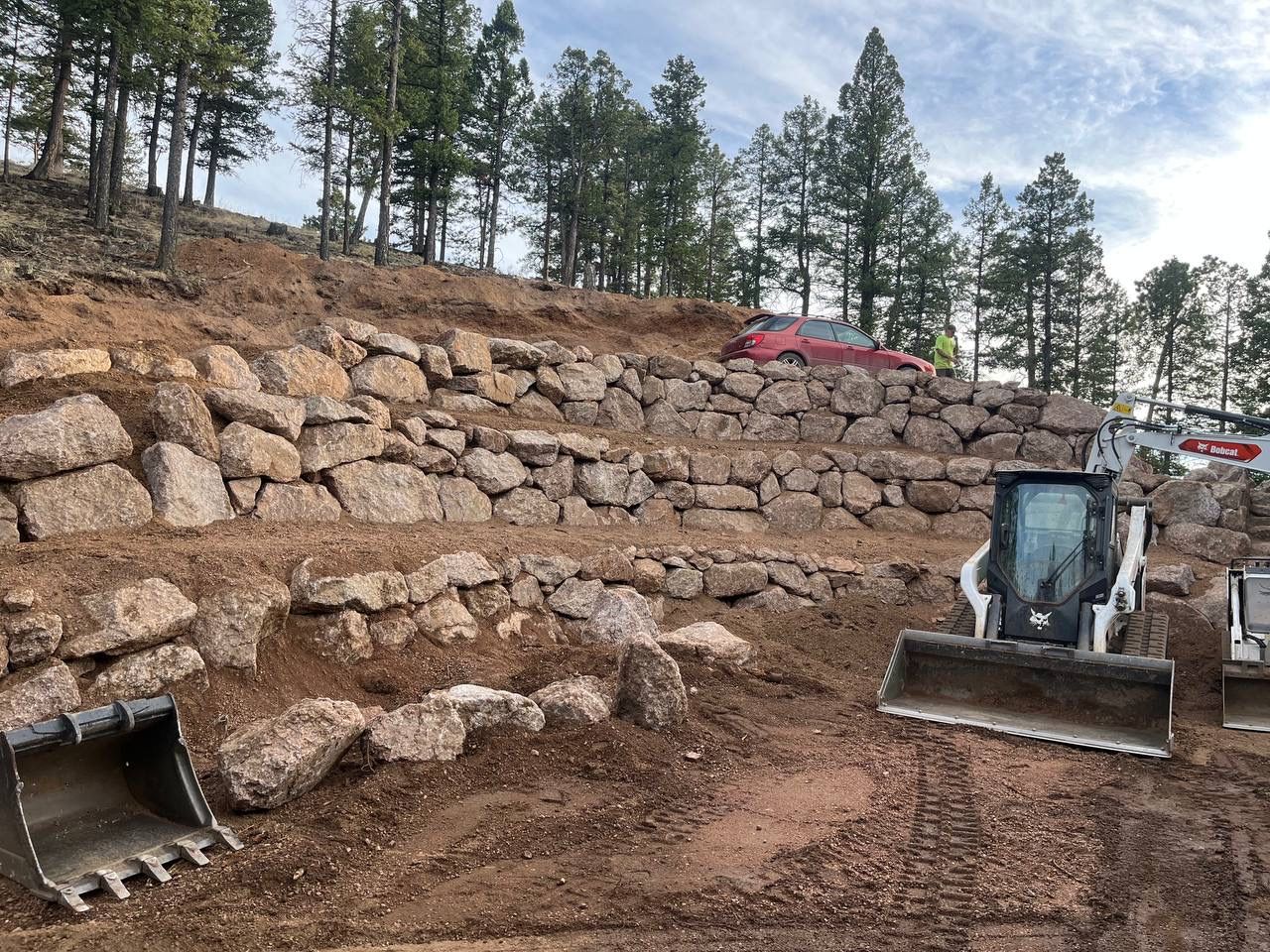 Excavator working on a terraced rock retaining wall at a wooded construction site