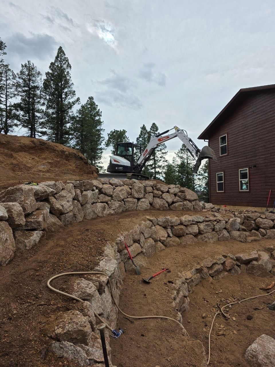 Excavator working on a terraced stone retaining wall beside a brown house in a wooded yard.