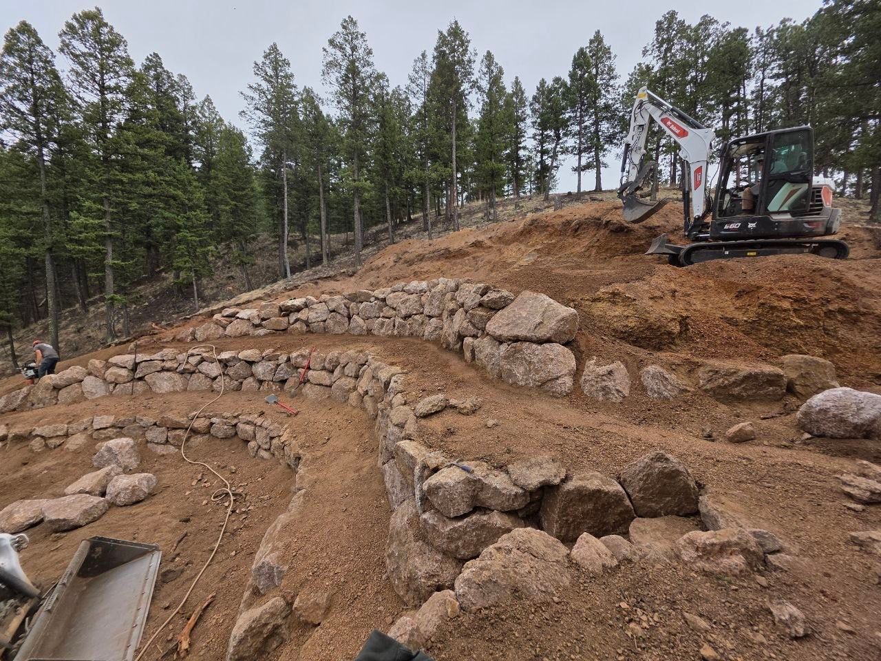 Excavator grading a rocky hillside with stone retaining walls in a pine forest.