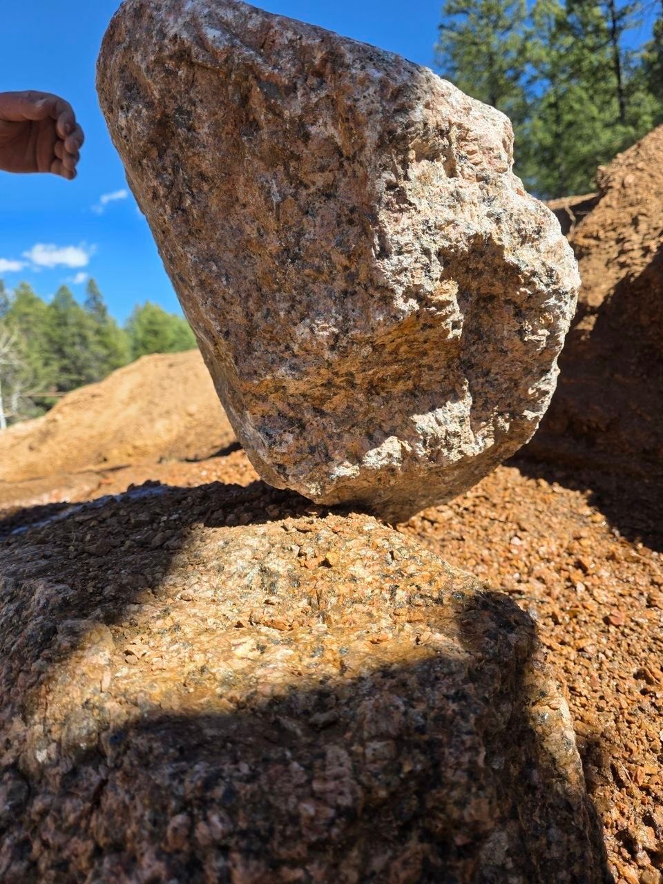 Large granite boulder balanced on a smaller rock under a clear blue sky in a wooded area