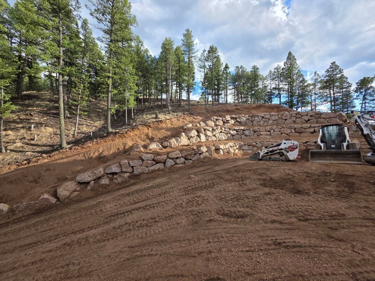 Rocky hillside under construction with excavator and dirt mound beside a pine forest.