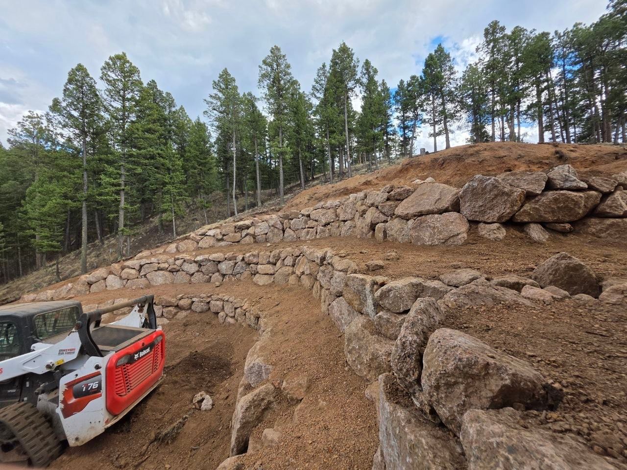 Construction equipment grading a rocky hillside beside a forest road