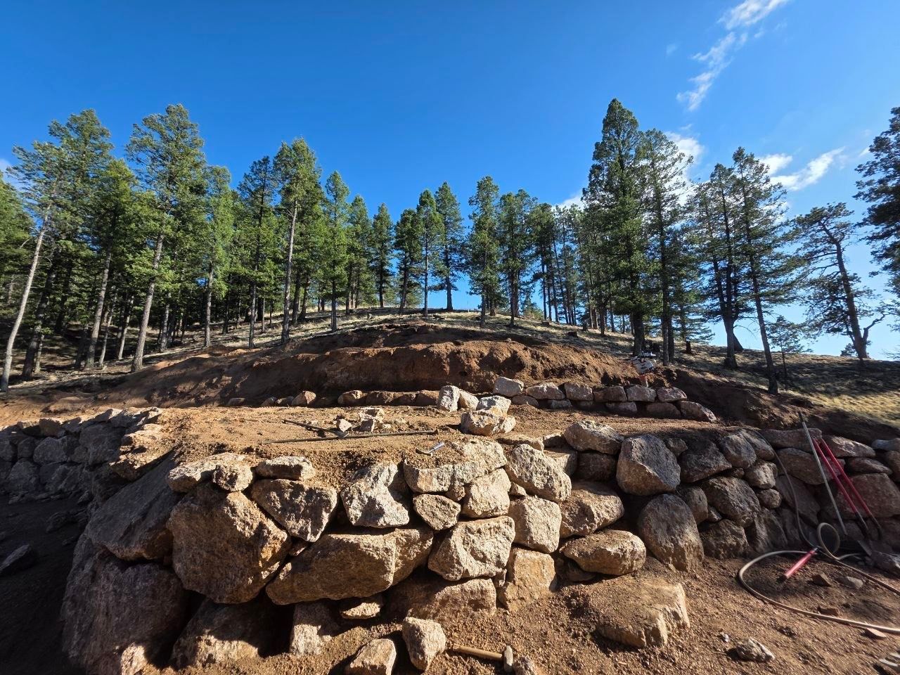 Stone retaining wall on a hillside with pine trees and blue sky above
