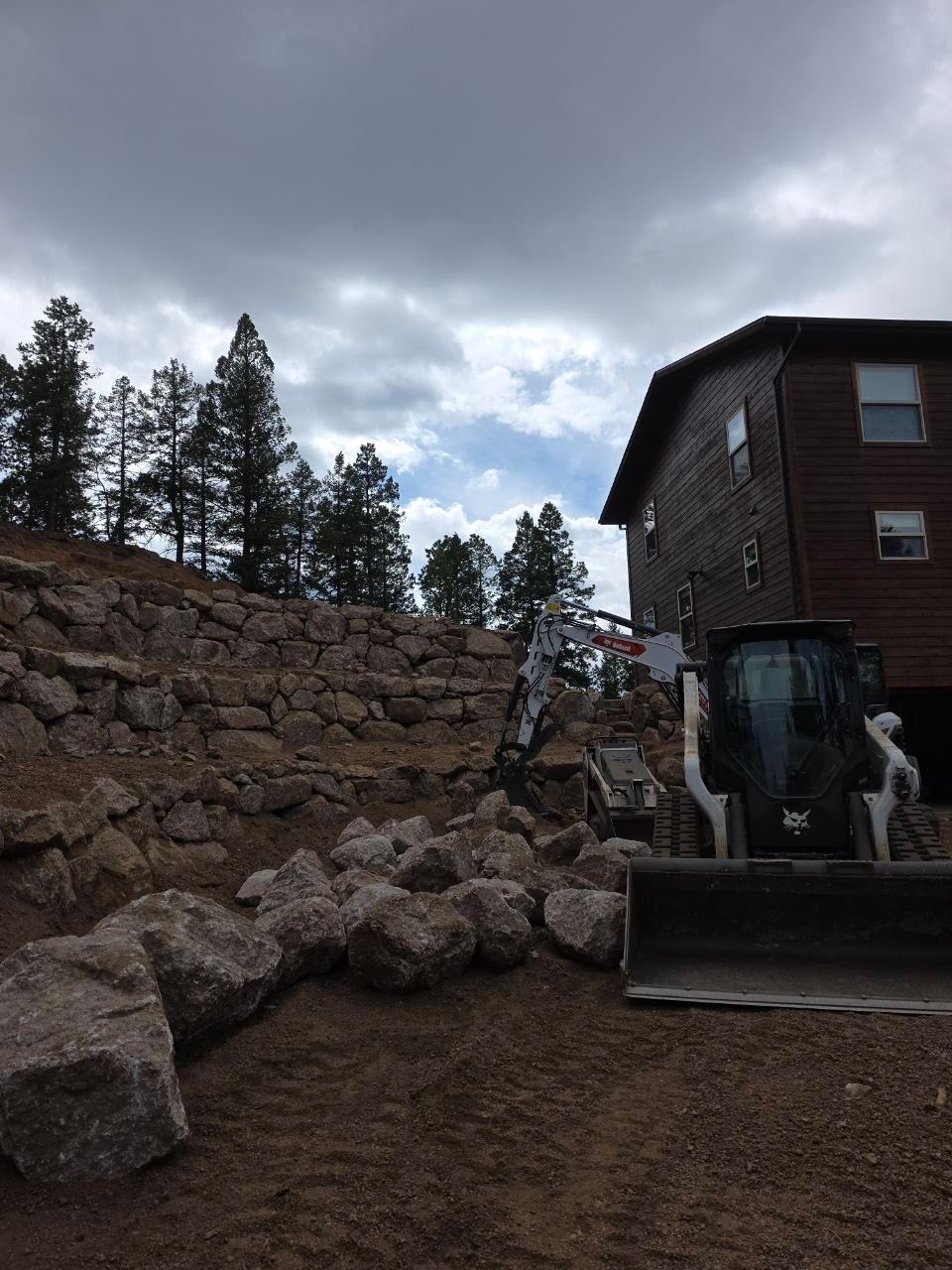 Construction equipment beside a stone retaining wall near a wooden house under a cloudy sky