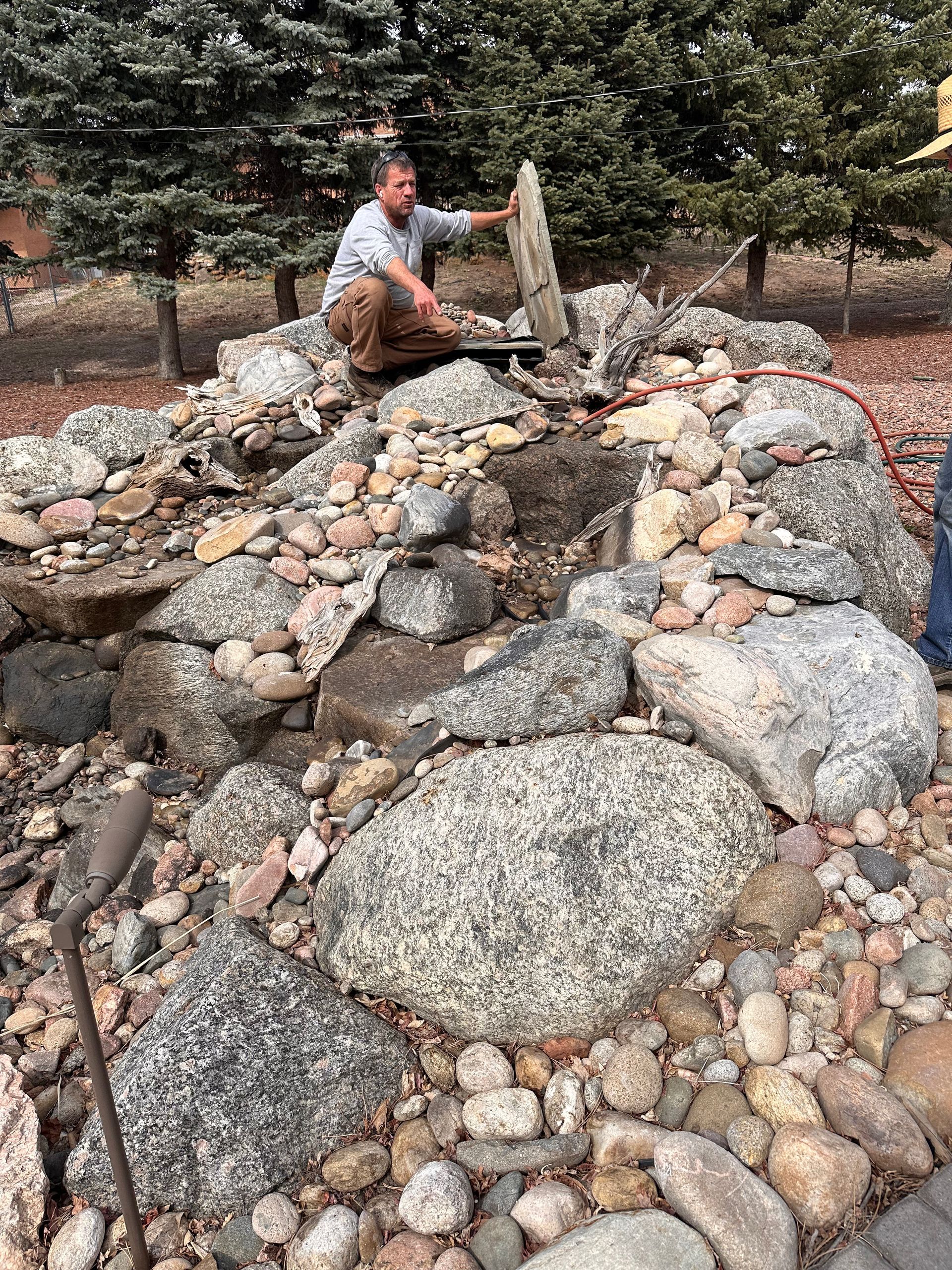 Person kneeling among large rocks in a landscaped creek bed with trees in the background