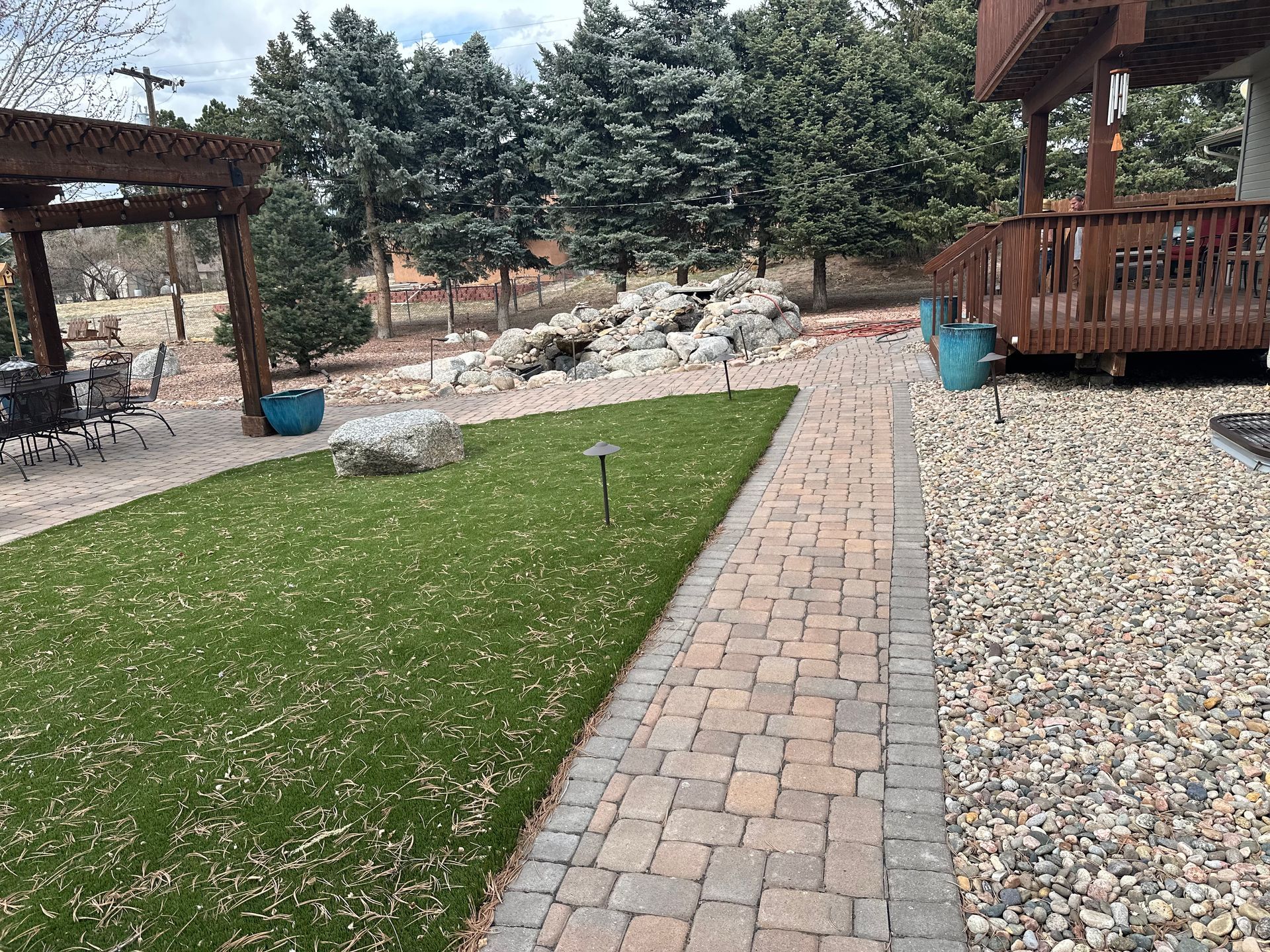 Backyard path beside green lawn, gravel, wooden deck, and pine trees under a pergola