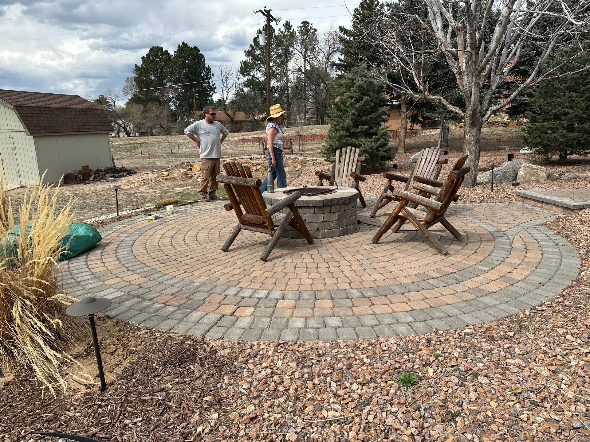 Two people standing by a circular stone patio with chairs and a fire pit in a wooded yard