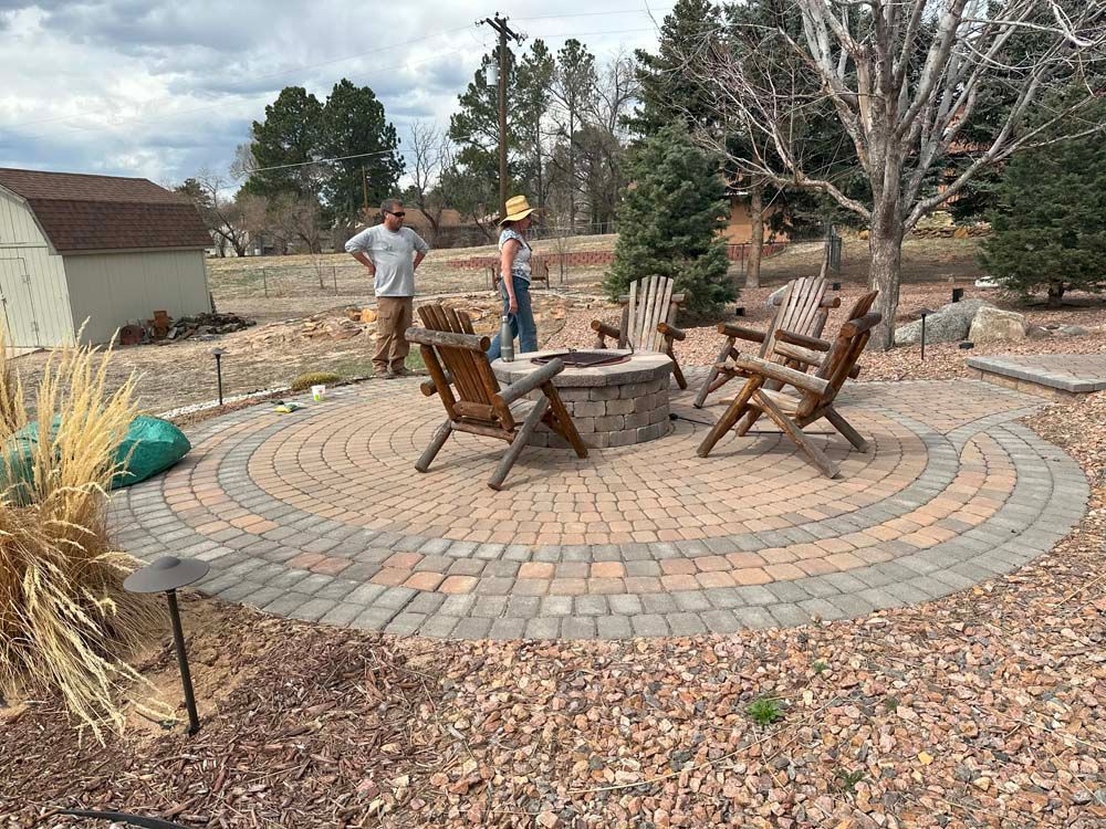 Circular stone patio with fire pit, wooden chairs, and two people standing nearby.