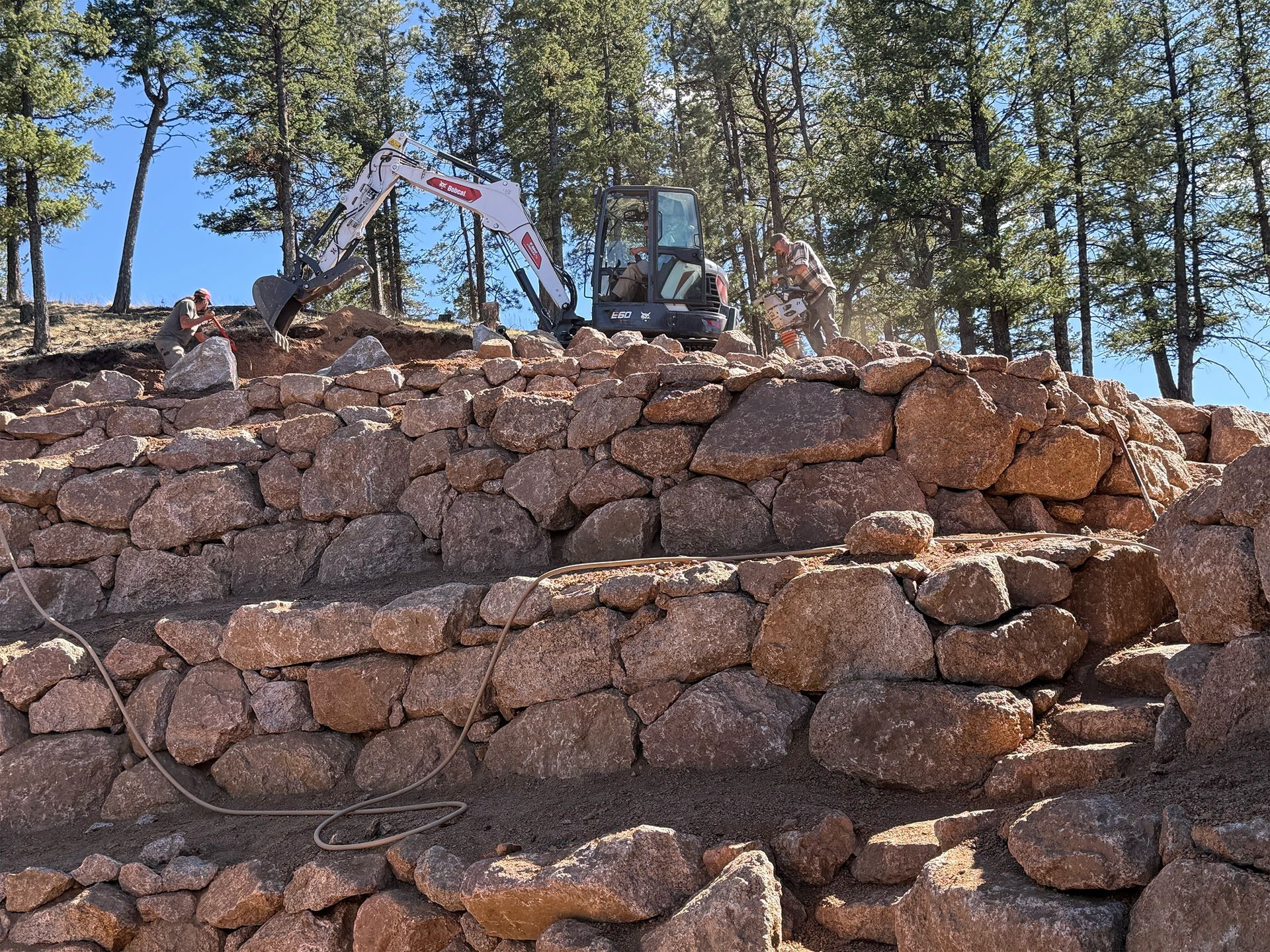 Stone retaining wall with a small outdoor machine and flagpole among pine trees under a blue sky