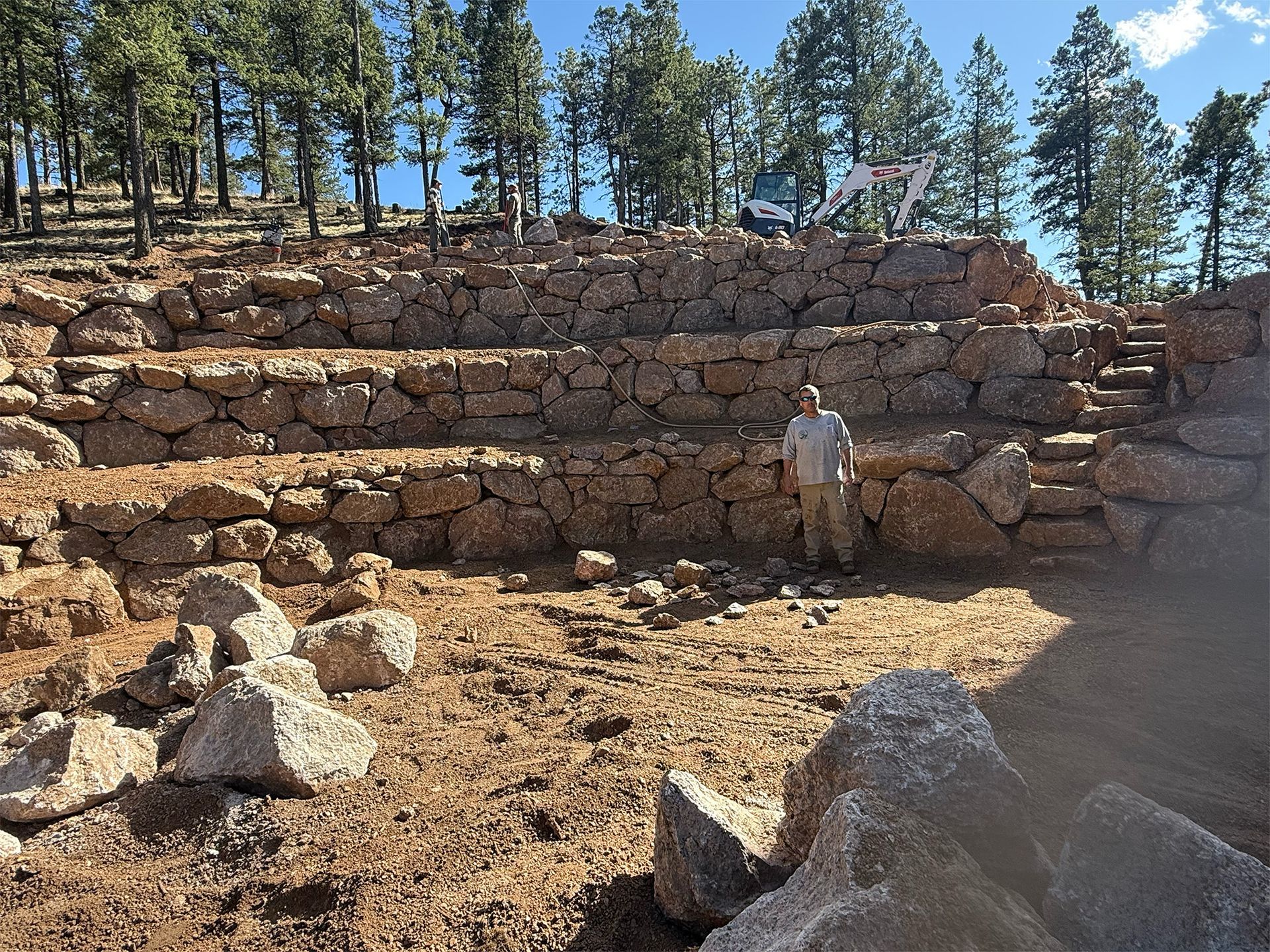 Terraced stone ruins on a hillside with a person standing near the center, trees in the background
