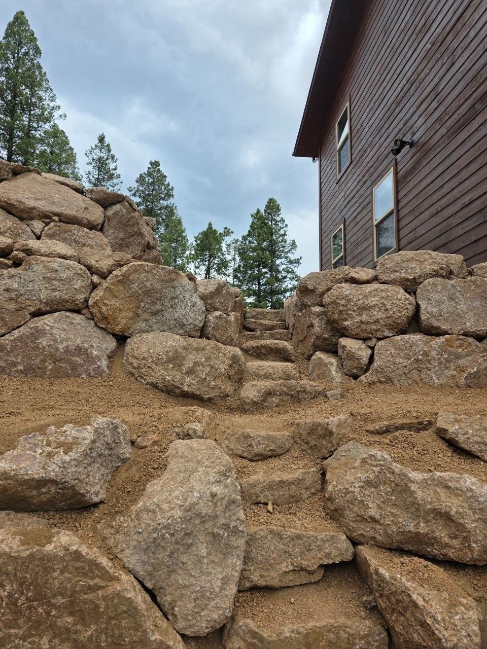 Rock retaining wall beside a brick house under a cloudy sky with pine trees in the background