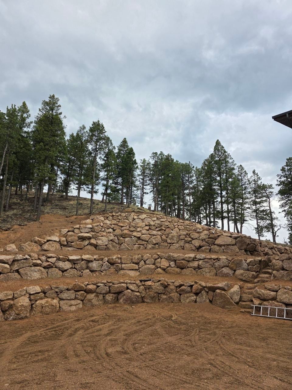 Stone retaining wall on a dirt hillside with pine trees under a cloudy sky
