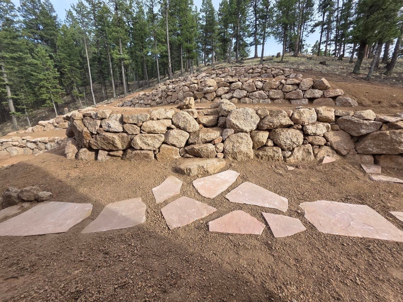 Stone retaining wall and flagstone path in a wooded outdoor area under construction.