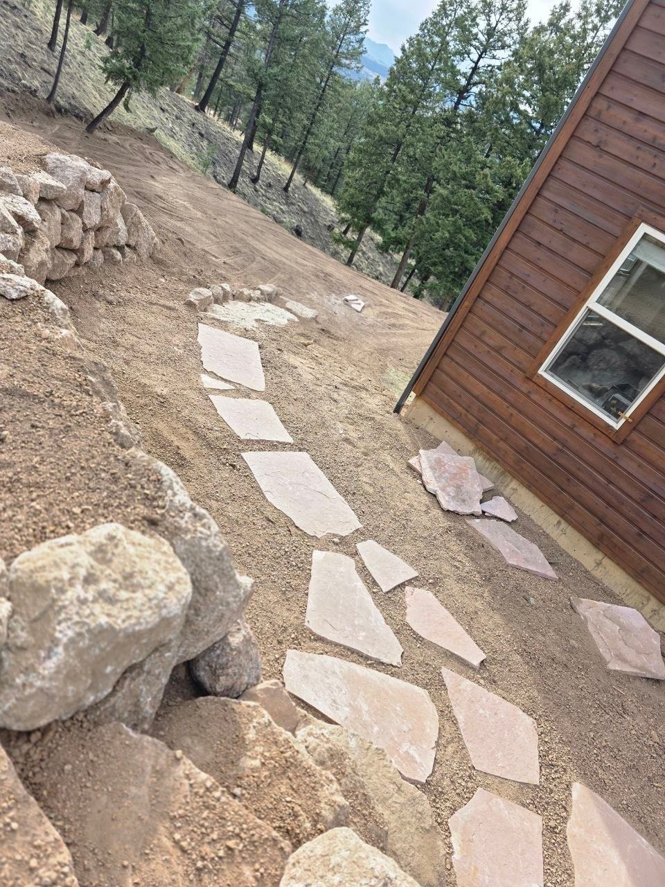 Stone path beside a brown cabin on a dry wooded hillside with rocks and trees