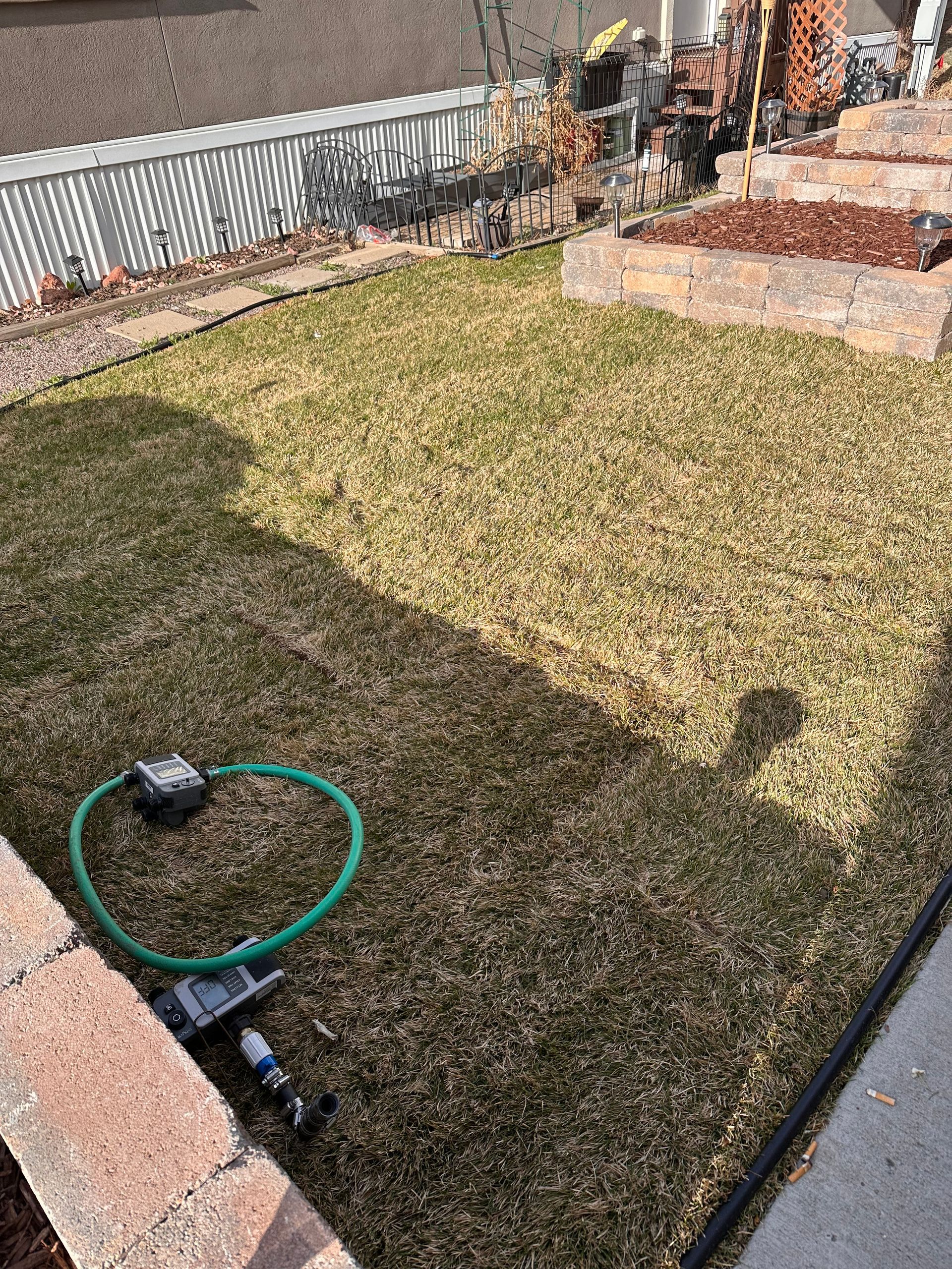 Backyard lawn with a green hose and sprinkler near a brick patio, viewed from above.
